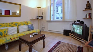 En stue med en gul sofa, et træsofabord, et fjernsyn på et stativ og et vindue med persienner.