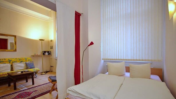Et soveværelse med seng, et natbord, en lampe, et vindue med persienner, en sofa og et lille bord.