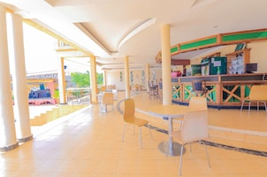 Um espaço amplo e coberto, com várias mesas e cadeiras, um balcão com diversos itens e vista para o exterior.