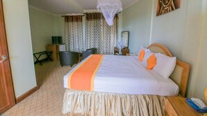Quarto de hotel com uma cama grande, uma mesa de cabeceira, uma cadeira, uma escrivaninha, abajur, ventilador e cortinas.