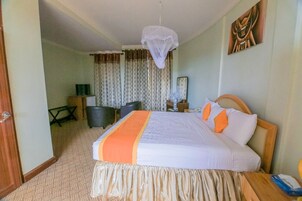 Quarto de hotel com uma cama grande, uma mesa de cabeceira, uma cadeira, uma escrivaninha, abajur, ventilador e cortinas.