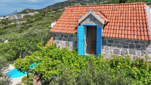 Una casa in pietra con tetto in tegole rosse e una porta blu, immersa nel verde con montagne sullo sfondo.