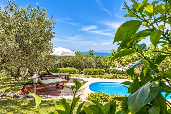 Un espace aménagé au bord de la piscine, avec des chaises longues et un parasol, entouré de verdure et offrant une vue sur la mer.