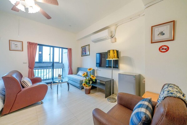 Una sala de estar con sofá, sillón y mesa de centro. Hay un televisor sobre un mueble bajo, una lámpara de pie y una ventana con cortinas.
