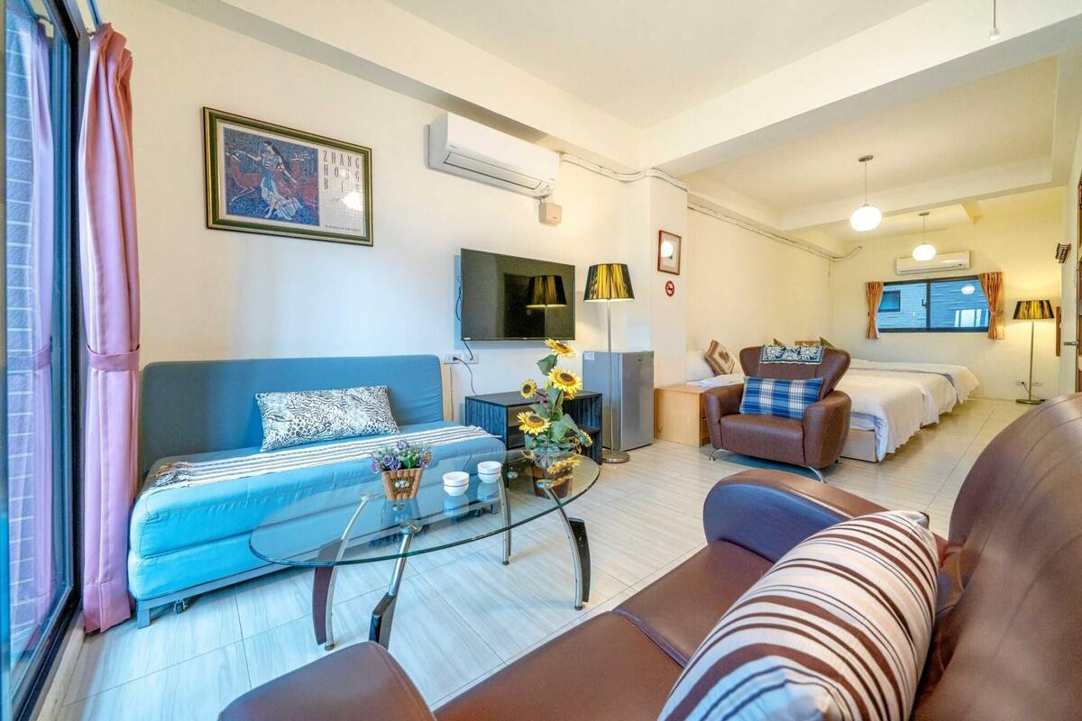 A modern living room with a blue sofa, a glass coffee table, and a brown leather armchair. There is a flat-screen TV mounted on the wall, a small table with a vase of flowers, and a bed with a blue and white striped bedspread in the background.