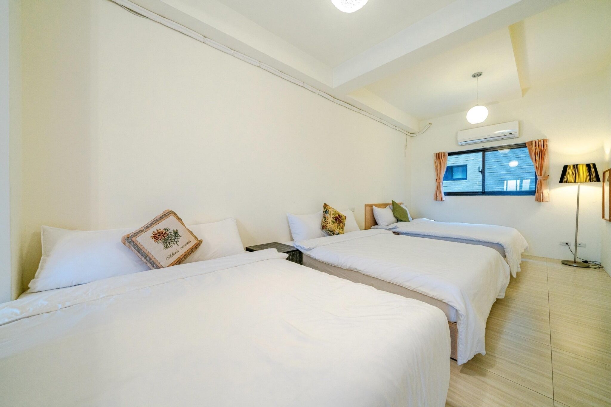 A hotel room with three beds, a window with curtains, an air conditioning unit, and a floor lamp.