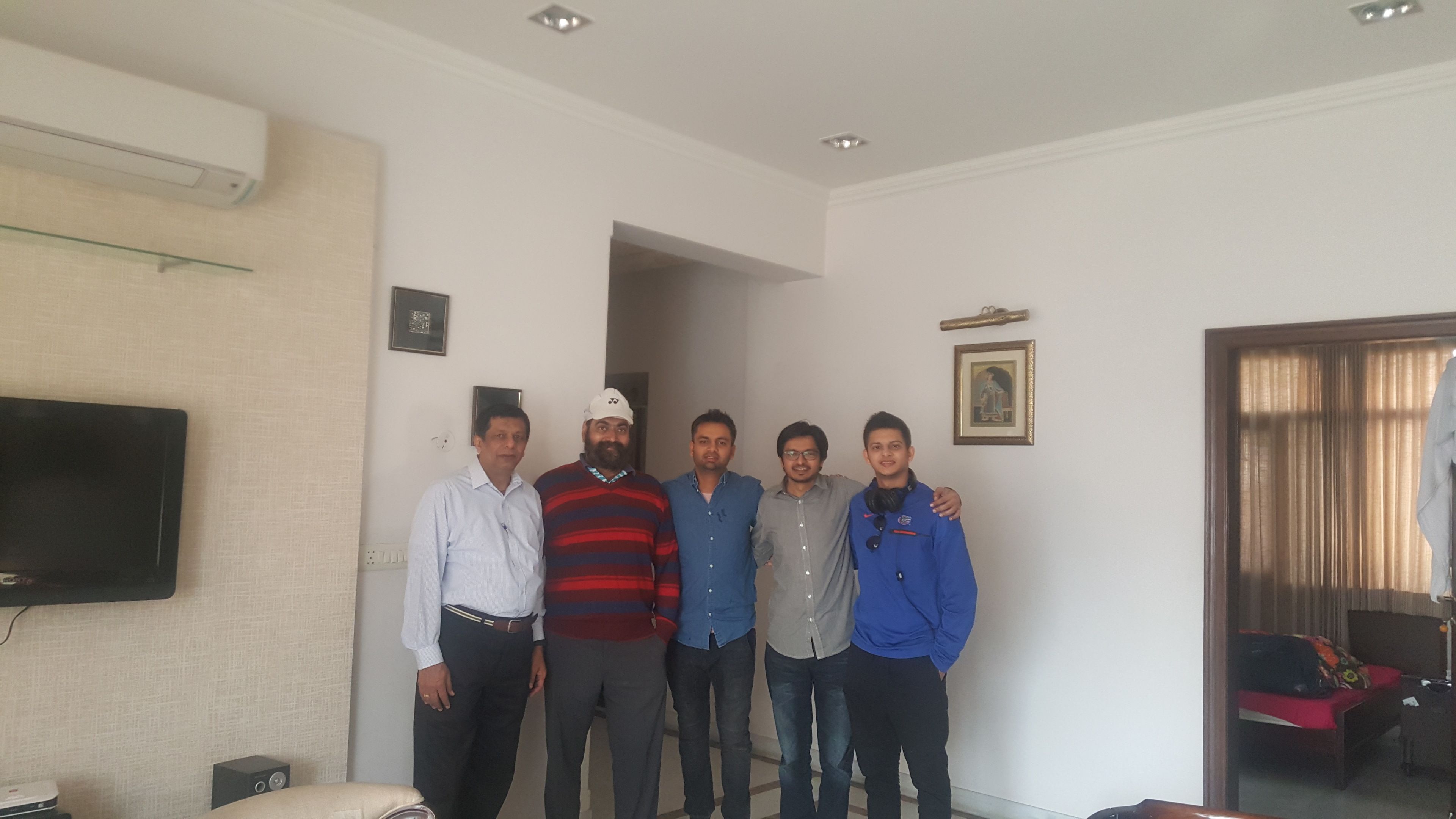 Un grupo de personas en una habitación con un televisor de pantalla plana montado en la pared, una cama con una colcha roja y una puerta que da a otra habitación.