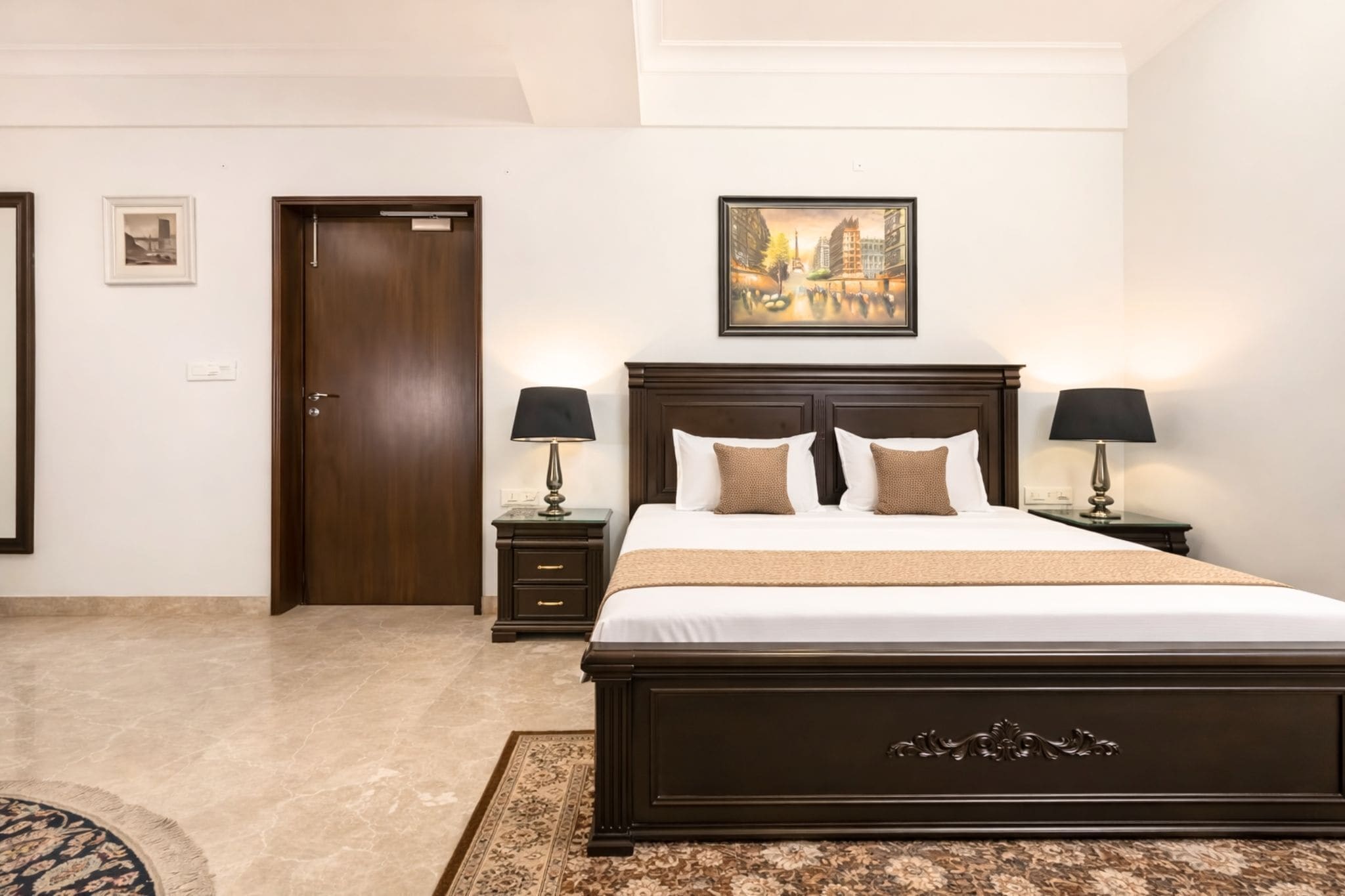 A hotel room with a large bed, two bedside tables with lamps, a door, a framed picture on the wall, and a patterned rug.