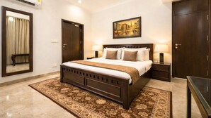 A bedroom with a large bed, a nightstand, a bedside table, a mirror, a door, and a painting on the wall. - Palm34 (Jaipur)