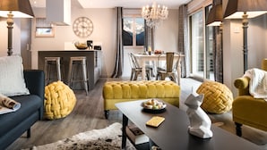 Una sala de estar moderna con sofá, reposapiés y mesa de centro, un comedor con una araña, y una cocina con un reloj y taburetes de bar.
