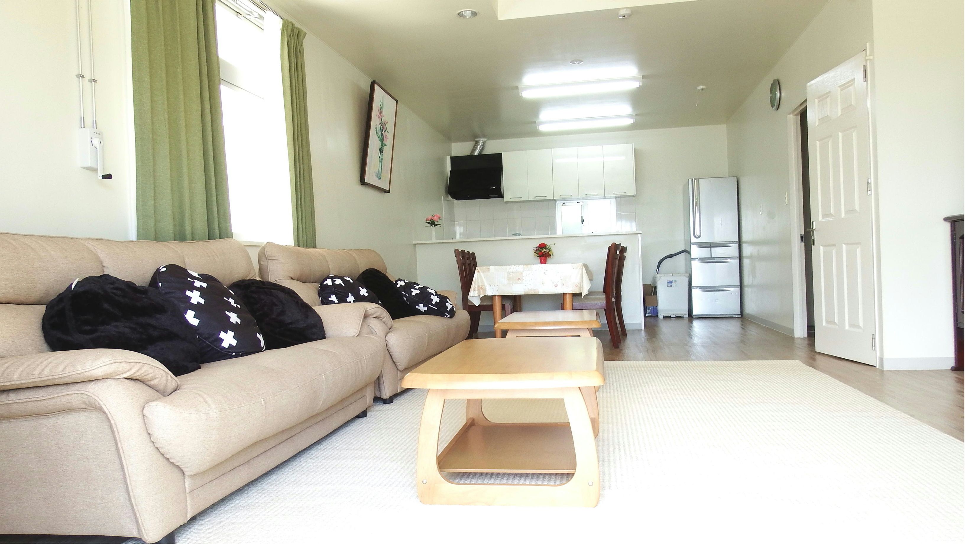 A living room with a beige sofa, a wooden coffee table, and a dining table with chairs. There is a kitchen area with white cabinets and a refrigerator.