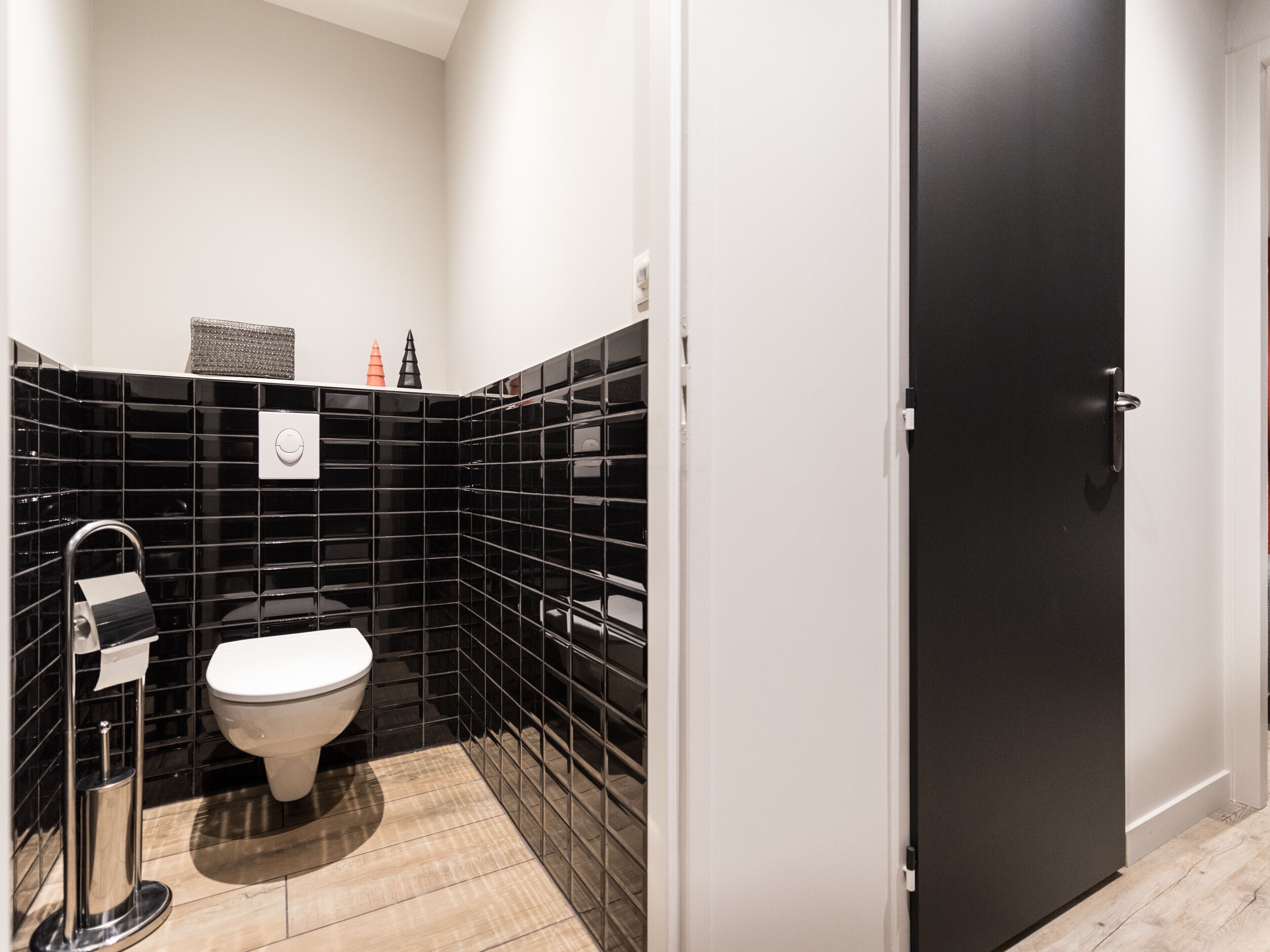 Baño pequeño con inodoro, paredes de azulejos negros y suelo de madera.