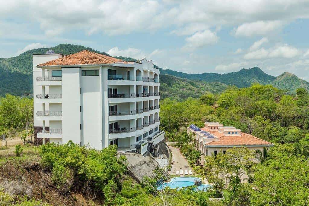 A multi-story hotel with balconies, surrounded by lush greenery and mountains.