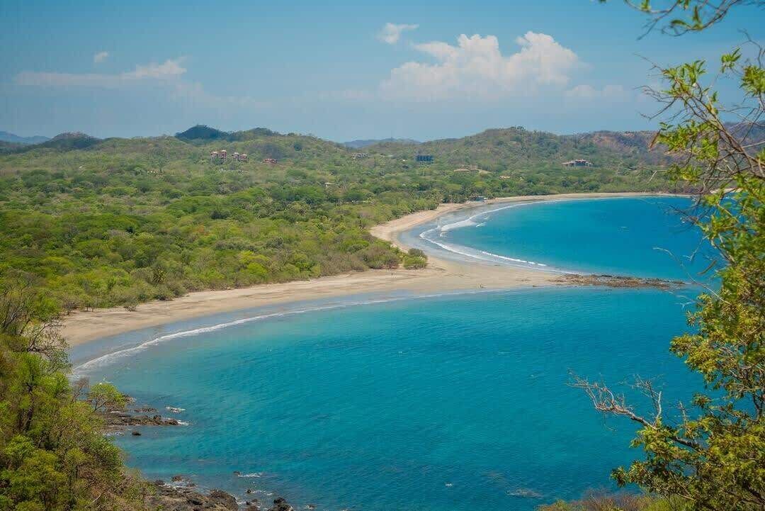 A coastal landscape with a beach, clear blue waters, and surrounding greenery.