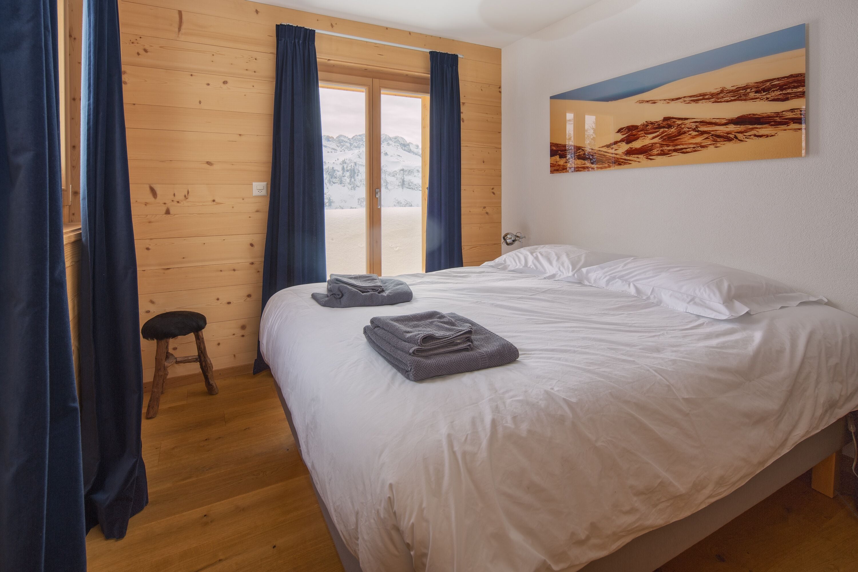 Une chambre avec un plancher en bois, un lit recouvert de draps blancs, un petit tabouret et une vue sur un paysage enneigé, visible à travers une porte coulissante.