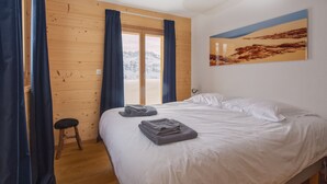 A bedroom with a wooden floor, a bed with white linens, a small stool, and a view of a snowy landscape through a sliding door. - eco-Chalet in Les Crosets (Valais)