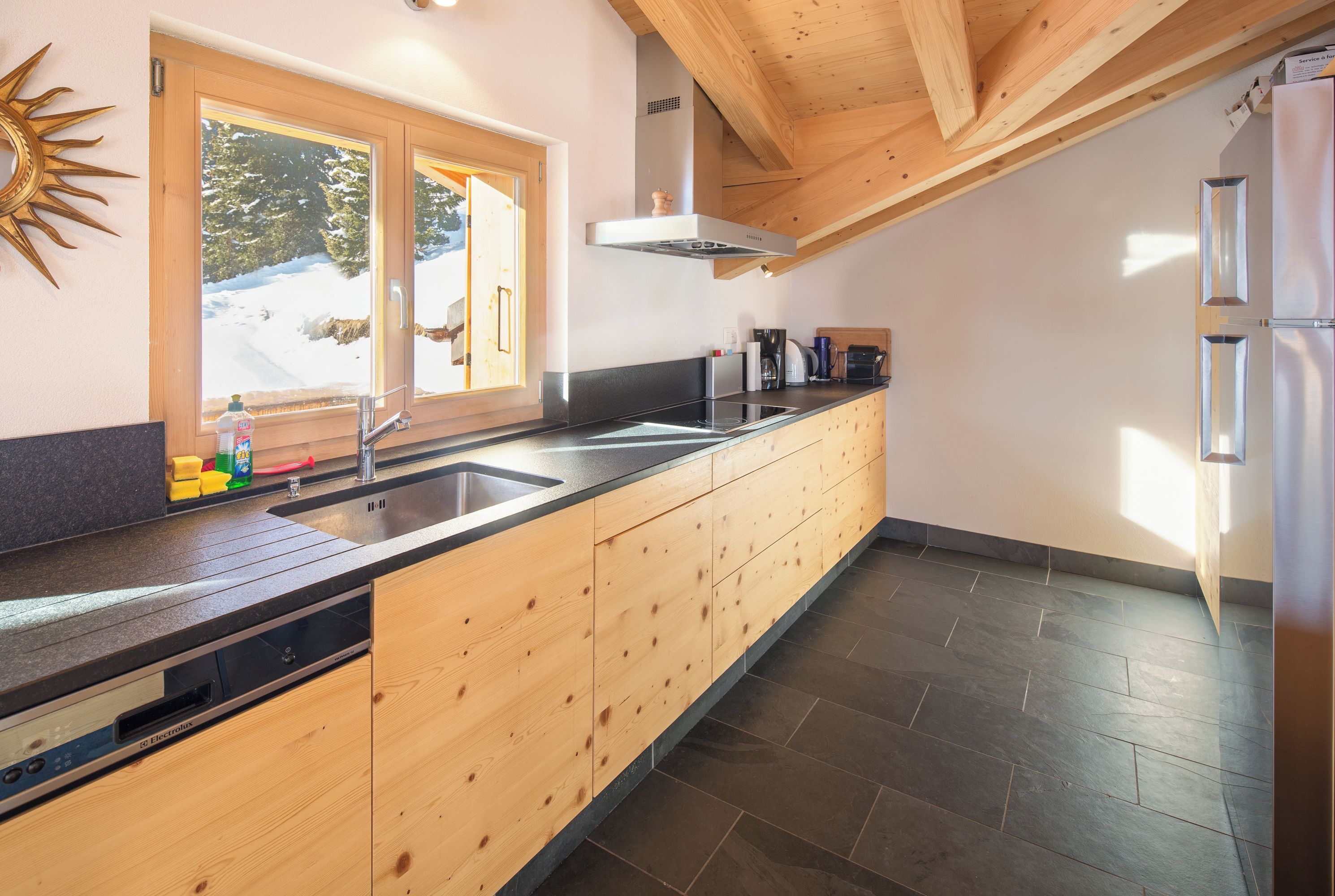 Eine moderne Küche mit Holzschränken, einer schwarzen Arbeitsplatte, einem eingebauten Geschirrspüler und einem großen Fenster mit Blick auf eine verschneite Landschaft.