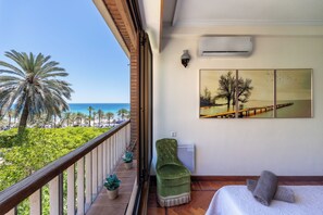 A balcony with a view of the sea and palm trees, a green chair, a bed with a white cover, and a painting on the wall.