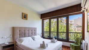 A bedroom with a large bed, a nightstand, a green armchair, and a window with a view of buildings and trees.
