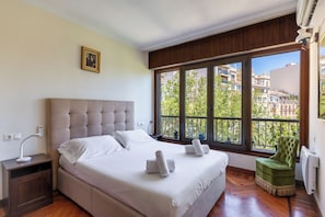 Une chambre à coucher avec un grand lit, une table de chevet, un fauteuil vert et une fenêtre donnant sur des immeubles et des arbres.