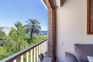 A balcony with a view of palm trees and the ocean, a grey sofa, and a hanging plant.