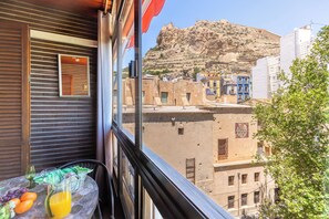 A balcony with a table set with fruit and a glass of orange juice, overlooking a cityscape with historic buildings and a prominent hill.