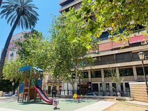 Un terrain de jeux avec un toboggan et des balançoires, un palmier et un bâtiment doté de balcons et d’une enseigne indiquant « Hôtel PASETTO ».