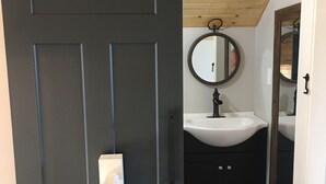 A dark wooden door leading to a bathroom with a white sink, a round mirror, and a wooden ceiling. - Ridge Cottage Residence (Campobello)