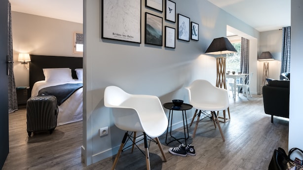 Una camera d'albergo moderna con un letto, una scrivania, due sedie bianche, un tavolino e una valigia.