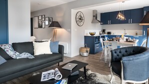 Un soggiorno moderno con un divano nero, un tavolino da caffè con riviste, una zona pranzo con un tavolo e sedie bianche e una cucina con mobili azzurri e un lampadario.