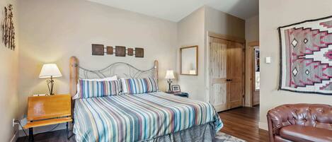 A bedroom with a bed, bedside lamps, a wooden bench, a patterned rug, a brown leather sofa, and a door leading to another room.