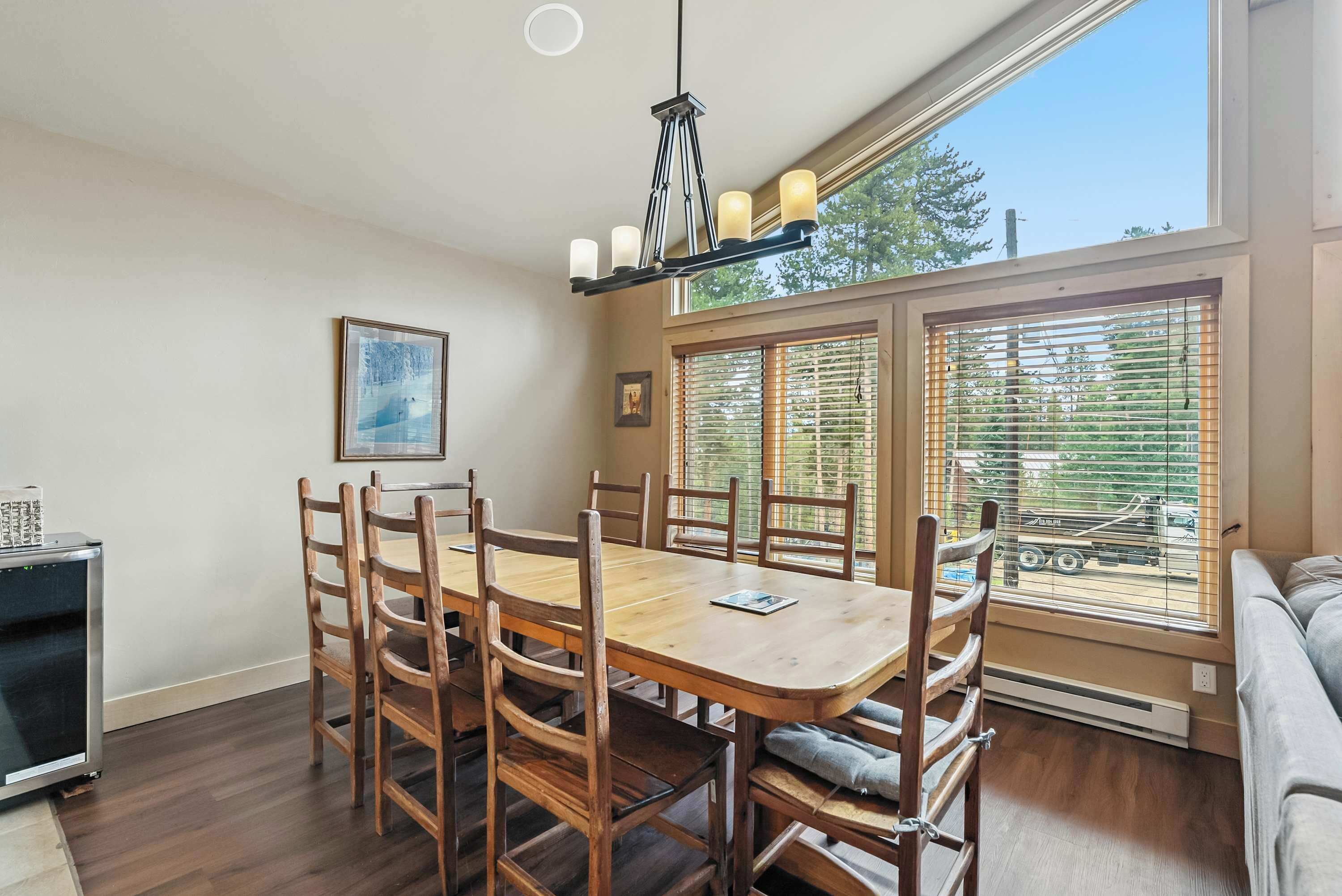 A dining area with a wooden table and chairs, a chandelier, and large windows with blinds.