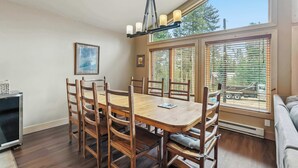 A dining area with a wooden table and chairs, a chandelier, and large windows with blinds.