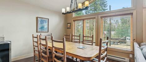 A dining area with a wooden table and chairs, a chandelier, and large windows with blinds.