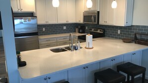 A modern kitchen with white cabinets, a stainless steel refrigerator, a microwave, and a sink. There are two pendant lights above the island and three stools. -  Beachfront, Beach Views, Great Location (Port St. Joe)