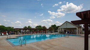 A swimming pool with lounge chairs and a covered area, surrounded by trees and a clear sky.