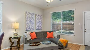 A living room with a grey sectional sofa, a glass coffee table, and a small side table with a lamp. There are two wall-mounted art pieces and a window with blinds.