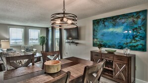 A dining area with a wooden table, chairs, a large abstract painting, and a modern pendant light. - Sunset Jetty View! Enjoy one free beach chair set up and umbrella each day of yo (Destin)