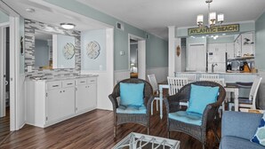 A spacious living area with a dining table, wicker chairs, and a kitchen area with white cabinets and a sign reading "ATLANTIC BEACH". - Direct OCEAN FRONT CONDO gorgeous view A Place at the Beach, Beautifully Updated (Atlantic Beach)