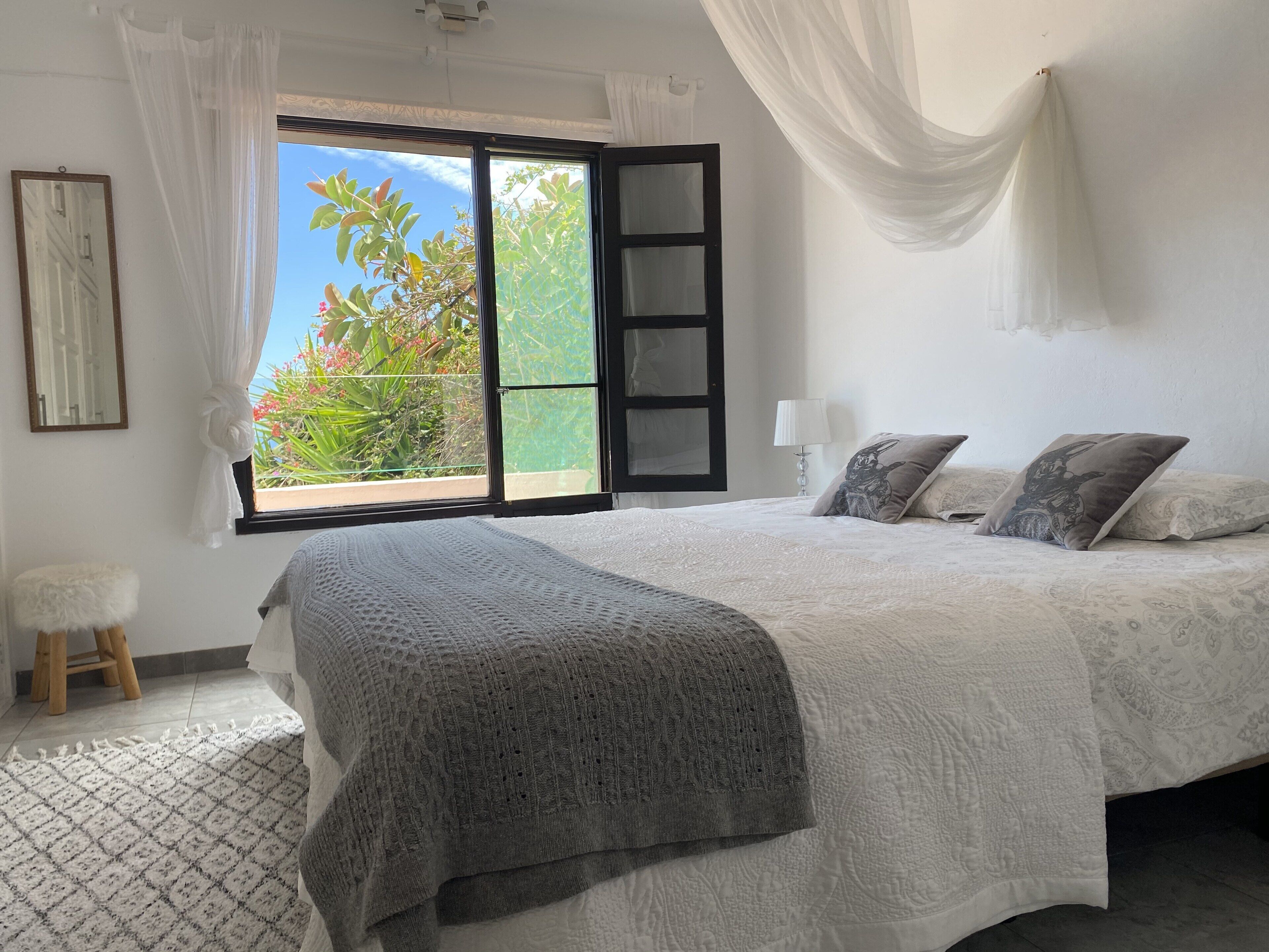 A bedroom with a large bed, a window with a view of greenery, a small stool, and a lamp.