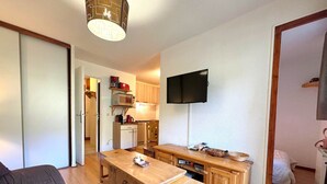 A compact, well-lit room with a kitchenette, a flat-screen TV mounted on the wall, a wooden dining table, and a bed with a dark-colored blanket.