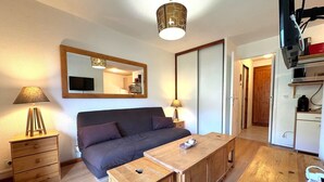 A compact bedroom with a large bed, a wooden nightstand, a bedside table, a mirror, and a kitchenette area.