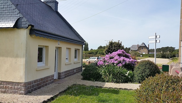 A two-story house with a beige exterior and dark roof, surrounded by a well-maintained garden with flowering plants and a gravel pathway.