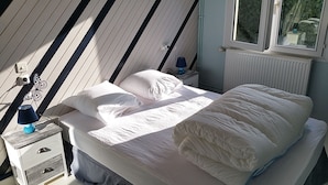 A bedroom with a sloped ceiling, a bed with white bedding, a nightstand with a lamp, and a window with a view of greenery.