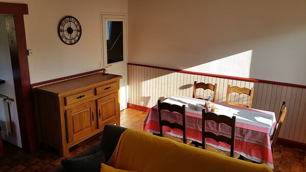 A room with a wooden dresser, a bed with a striped bedspread, a dining table with chairs, a clock on the wall, and a window with blinds. - Perros-Guirec Ploumanac'h - house 6 people (Perros-Guirec)