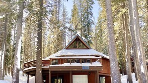A wooden cabin with a covered porch and a garage, surrounded by tall trees and covered in snow. - Lakeview Capone Getaway, great sledding on property, fun loft for kids, Lg deck (Soda Springs)