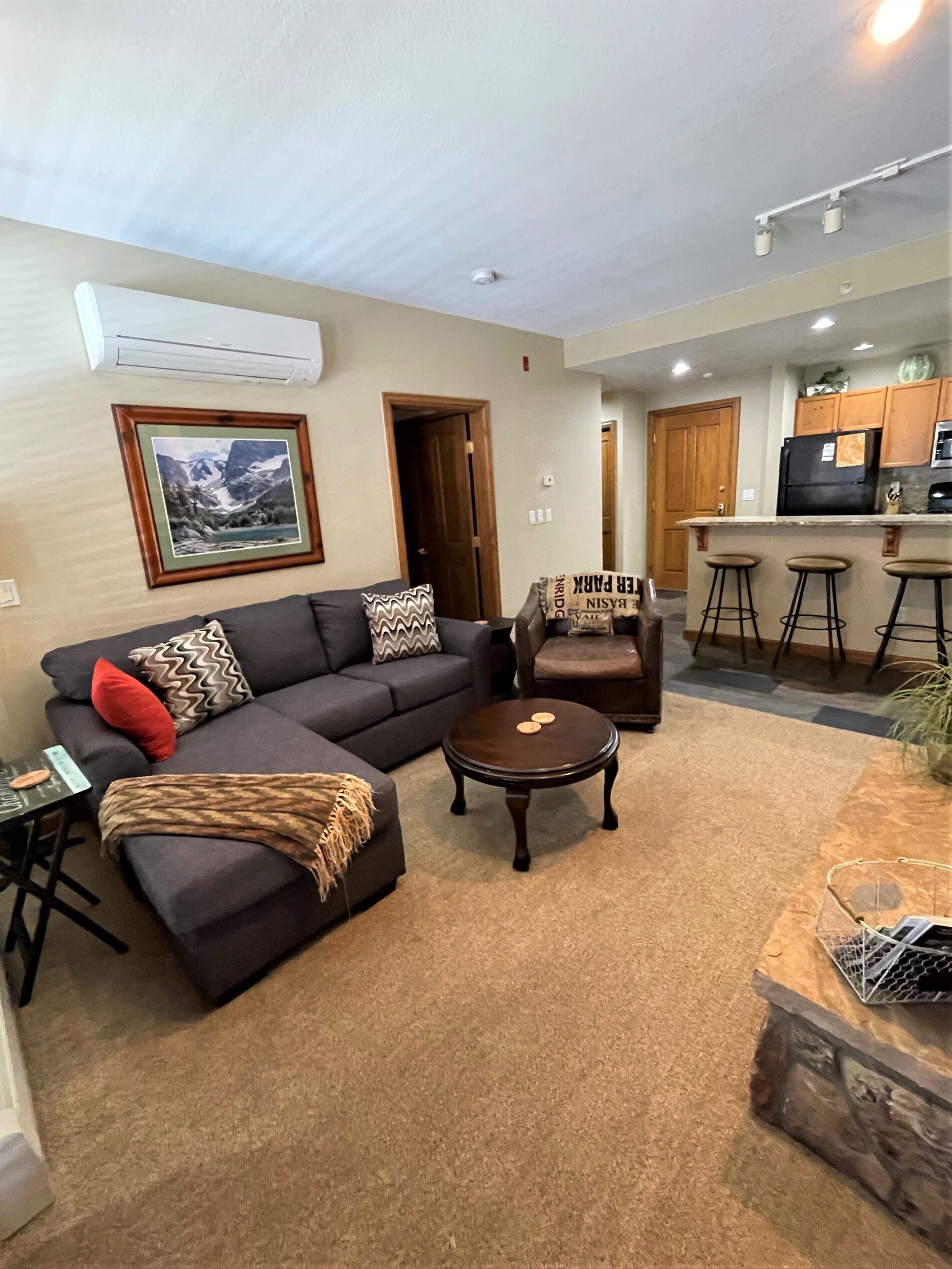 A living room with a sofa, armchair, and coffee table, a kitchen area with a refrigerator and bar stools, and a framed picture on the wall.