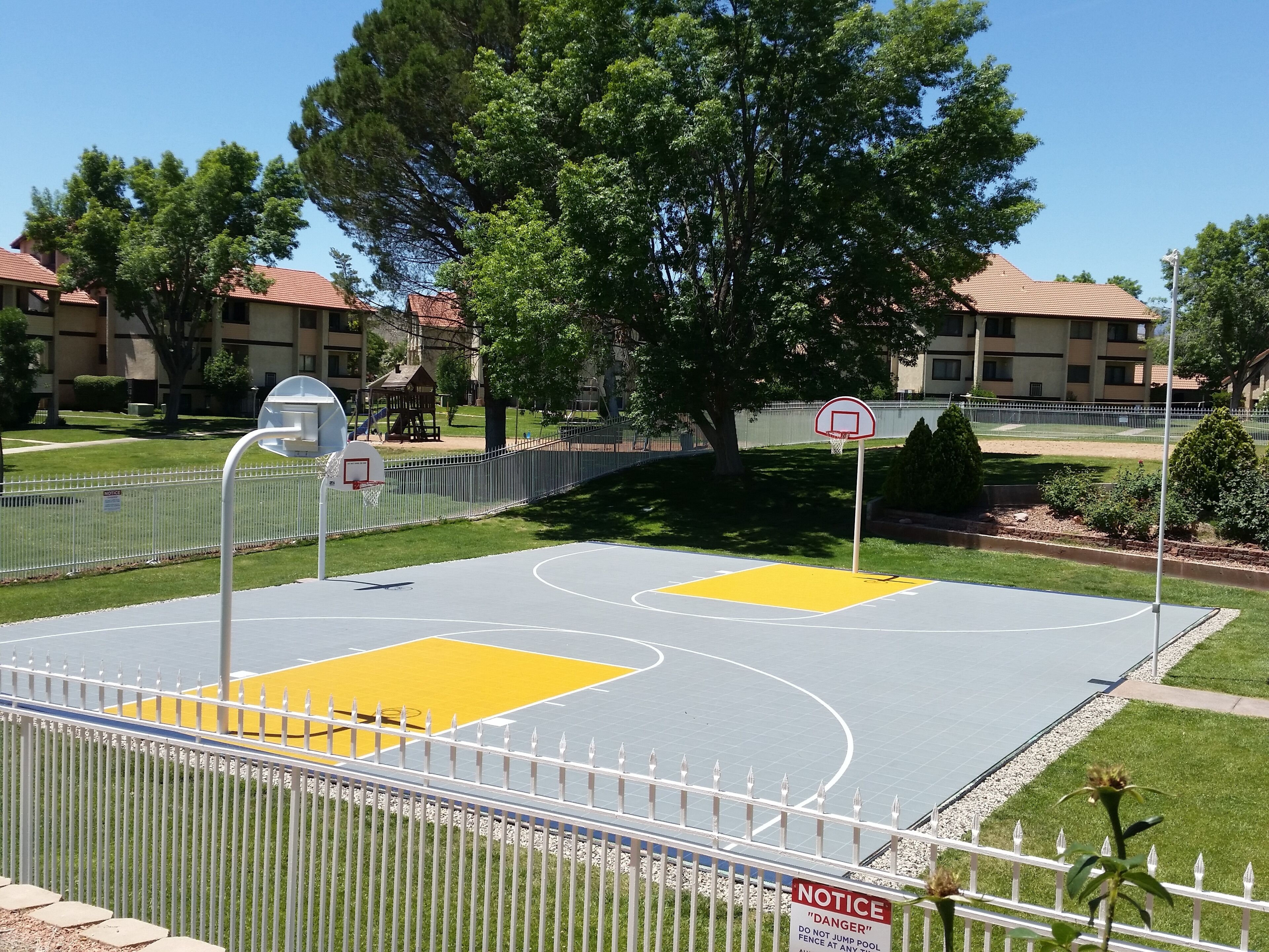 Un terrain de basket en plein air, entouré d’une clôture blanche et agrémenté d’un panneau d’affichage.
