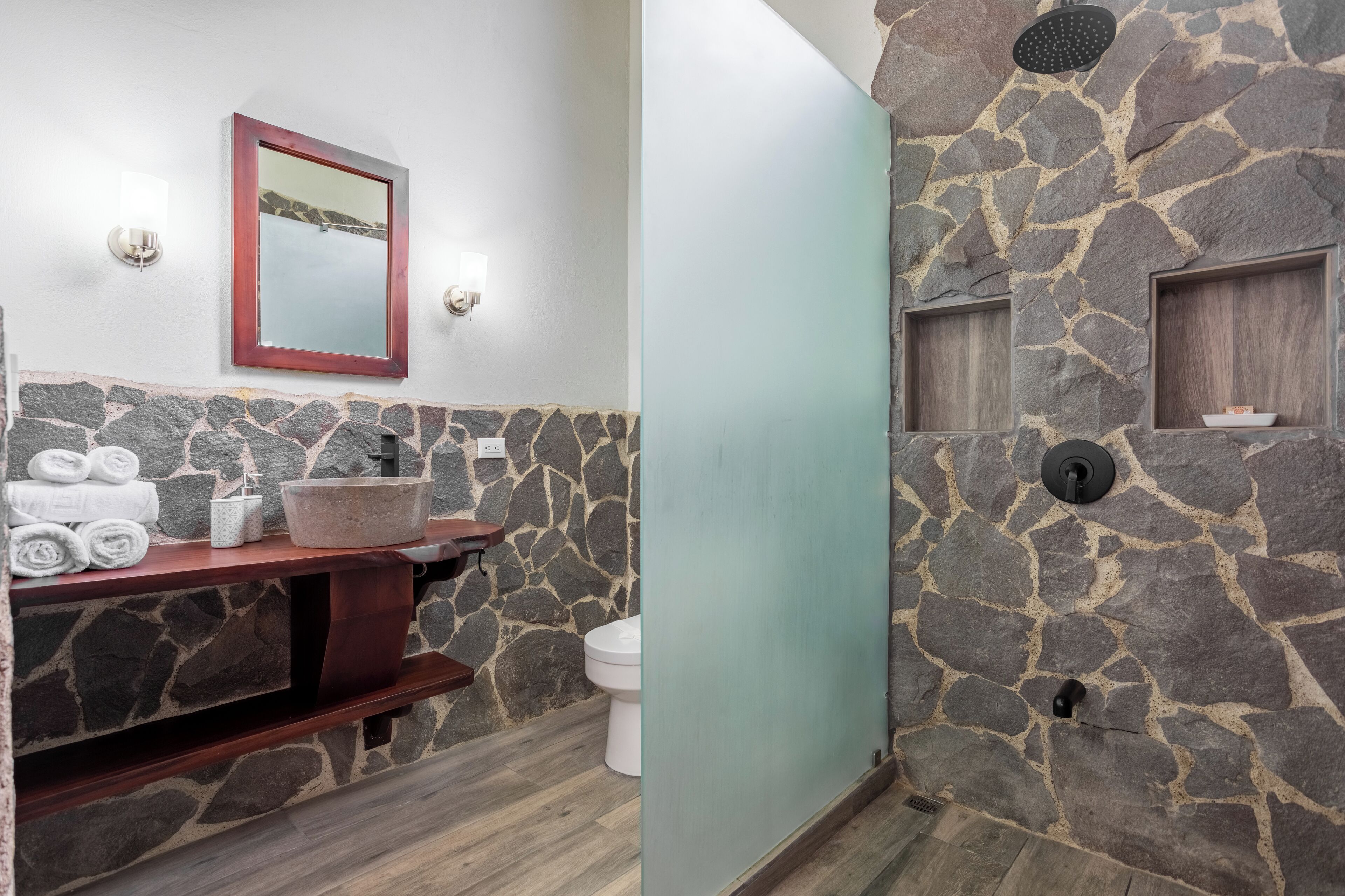A bathroom with a stone wall, a wooden vanity, a mirror, and a toilet.