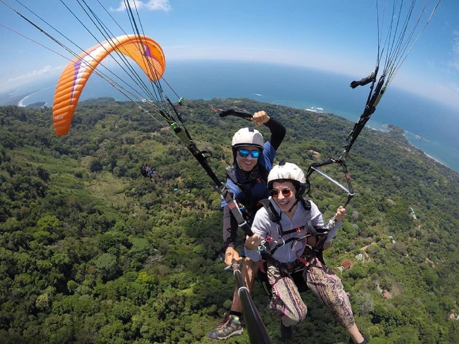 Dues persones fent paraglís sobre un paisatge verd i abundant amb vista a l'oceà.